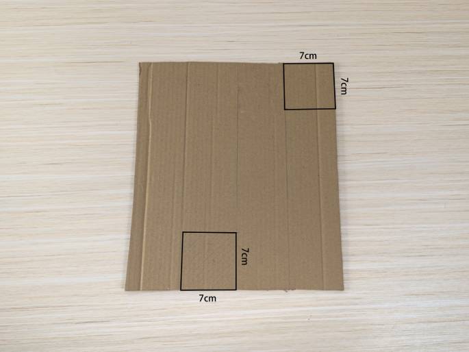 A piece of brown cardboard lies on a light wood-grain table. Two squares, measuring 7cm x 7cm each, are outlined on the cardboard. One square is located in the bottom left corner, and the other is in the top right corner. The cardboard appears to be marked for cutting or folding along these lines.