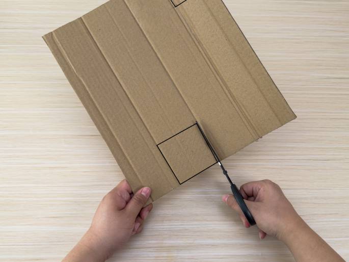 A pair of hands is cutting a piece of brown cardboard with scissors. The cardboard, placed on a light wood-grain table, has a square outlined on it, and the scissors are cutting along one side of this square. The left hand holds the cardboard steady, while the right hand operates the scissors.