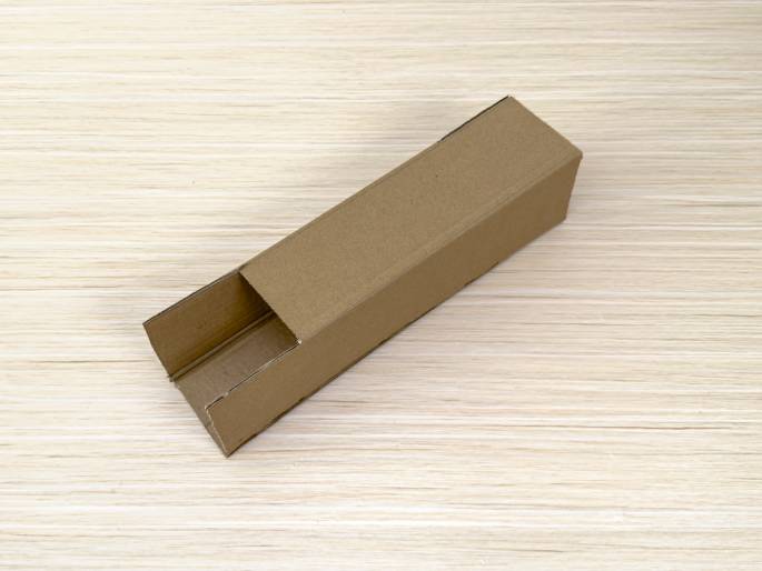 A long, rectangular box made of brown cardboard sits diagonally on a light wood-grain table. One end of the box is open, revealing the hollow interior, while the other end is closed. The box appears to be partially constructed or deconstructed, with the open end showing folded cardboard flaps.