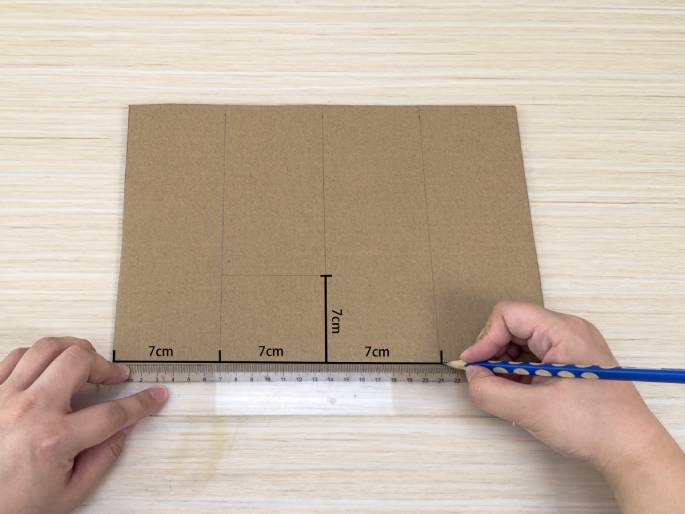 On a light wood-grain table, a piece of brown cardboard has been marked with lines and measurements. Two hands are shown measuring and marking the cardboard. One hand holds a clear plastic ruler along the bottom edge, while the other hand uses a pencil to draw a line. Three 7cm markings are visible along the bottom edge, and a single 7cm marking is visible along a vertical line. The markings suggest the cardboard is being prepared for cutting or folding.