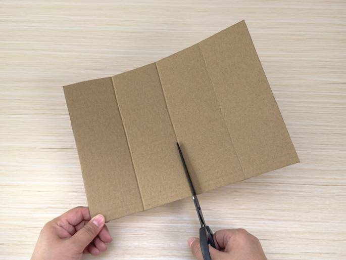 Two hands are shown cutting a piece of brown cardboard with scissors. The cardboard, which rests on a light wood-grain table, is folded slightly and has several vertical creases. The left hand holds the cardboard, while the right hand uses the scissors to cut along one of the creases.