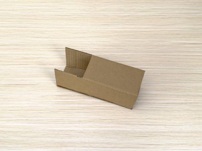 A small, open-ended cardboard box lies diagonally on a light wood-grain surface. The box is rectangular and made of plain brown cardboard. One end is fully open, while the other remains closed. The open end has folded flaps, suggesting the box is either partially assembled or disassembled.
