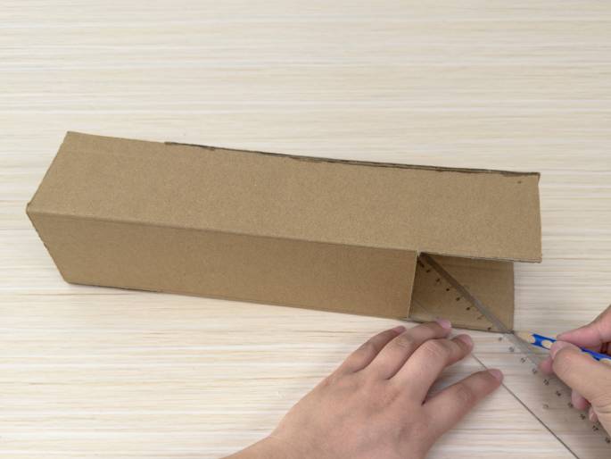 A partially assembled cardboard box sits on a light wood-grain table. Two hands are in the process of marking a line on one of the box's flaps. One hand holds a clear plastic ruler against the cardboard, while the other hand uses a pencil to draw along the ruler's edge. The box is long and rectangular, made of plain brown cardboard. One end is open, revealing the interior.
