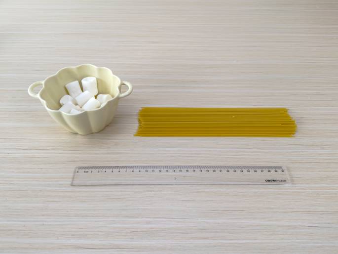 A small, light-yellow bowl filled with white marshmallows, a bundle of uncooked spaghetti, and a clear plastic ruler are arranged on a light wood-grain table. The items appear to be set up for a learning activity or a demonstration, possibly involving measurement or construction.