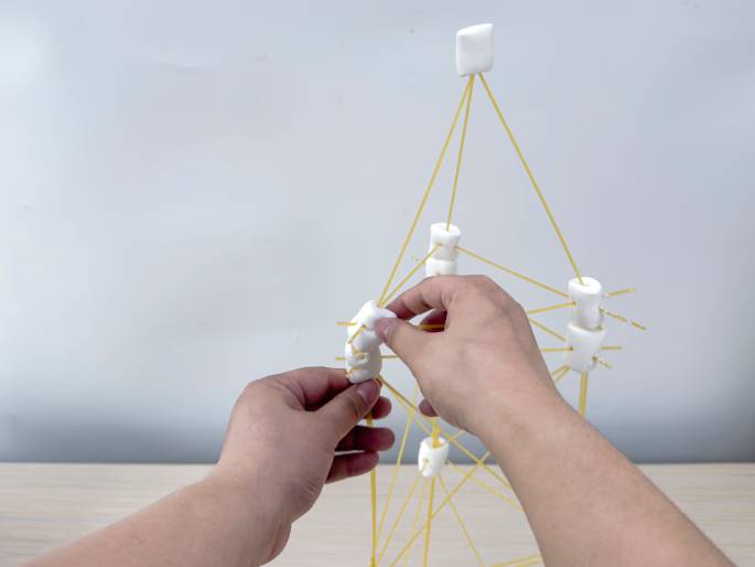 Two hands are in the process of building a tower-like structure with large white marshmallows and strands of uncooked spaghetti against a light-colored background. The marshmallows act as connectors, and the spaghetti pieces are inserted into them to create the framework. The structure appears to be a complex geometric design, and the hands are actively working to add more marshmallows and spaghetti, indicating ongoing construction. This image likely depicts a STEM activity, engineering project, or learning exercise.