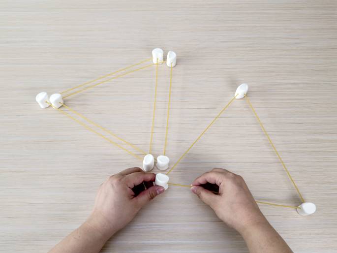 Two hands are constructing a geometric shape on a light wood-grain surface using large white marshmallows and thin, light-yellow rubber bands. The marshmallows serve as nodes, and the rubber bands connect them, forming the sides of what appears to be a series of connected triangles. The hands are in the process of adding another rubber band, suggesting the structure is still being built. This image likely depicts a STEM activity or a learning exercise.