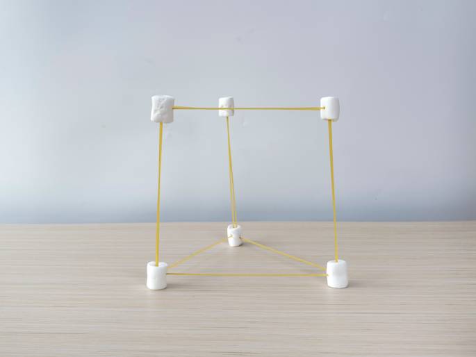 A three-dimensional structure made of large white marshmallows and uncooked spaghetti stands on a light wood-grain table against a plain off-white background. The base is a triangle formed by marshmallows connected with spaghetti. Vertical spaghetti pieces rise from these base marshmallows to connect with another set of marshmallows, creating a square or rectangular prism shape. This image likely depicts a STEM activity or educational project demonstrating structural principles.