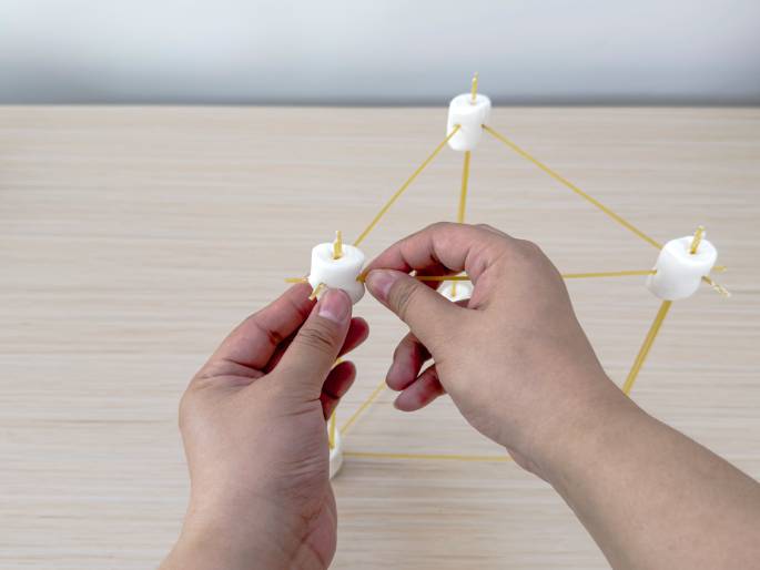 Two hands are in the process of building a structure with large white marshmallows and sticks of uncooked spaghetti on a light wood-grain table. The marshmallows act as connectors, and the spaghetti pieces form the struts of a partially completed geometric form. The hands are inserting a piece of spaghetti into a marshmallow, indicating the construction is in progress. This scene likely represents a STEM building activity or a learning game.