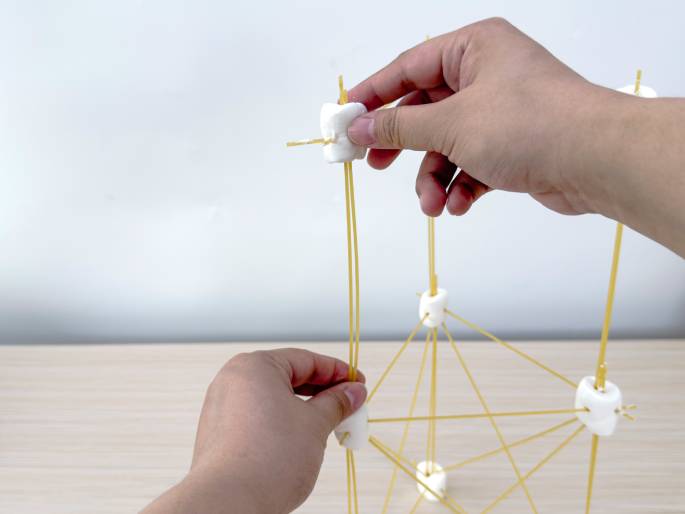 Two hands are building a structure with large white marshmallows and sticks of dry spaghetti on a light-colored wooden surface. The marshmallows are used as connectors, with spaghetti pieces inserted into them to create a framework. The structure appears to be a geometric shape, possibly a cube or prism, and is in the process of being assembled. The hands are actively adding more spaghetti pieces to the marshmallows, indicating ongoing construction. The image likely depicts an educational activity or a STEM project.