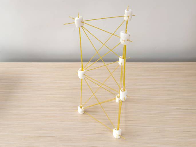 A freestanding structure made of large white marshmallows and uncooked spaghetti sits on a light wood-grain table against a plain off-white background. The marshmallows serve as connectors, with multiple spaghetti strands inserted into them, forming a tall, somewhat leaning tower or prism-like shape. The numerous spaghetti pieces create a complex lattice structure. This image likely represents a completed STEM building project or educational model.