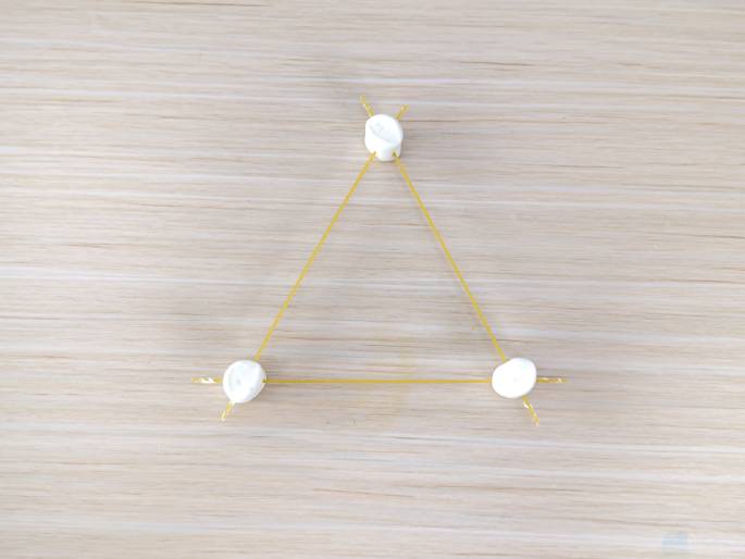 A simple triangular structure made of large white marshmallows and uncooked spaghetti lies flat on a light wood-grain table. Three marshmallows form the corners of the triangle, connected by pieces of spaghetti. Short, broken pieces of spaghetti protrude from the marshmallows, suggesting previous construction or attempts to connect more pieces. The image likely represents the base or a component of a larger STEM building activity.