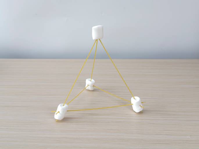 A tetrahedron-shaped structure constructed from large white marshmallows and uncooked spaghetti stands on a light wood-grain table against a plain, light-colored background. Three marshmallows form the base, connected by spaghetti pieces, and a fourth marshmallow sits atop the structure, also connected with spaghetti struts. Small, broken pieces of spaghetti stick out from the marshmallows. This image likely represents a completed STEM engineering or building challenge.