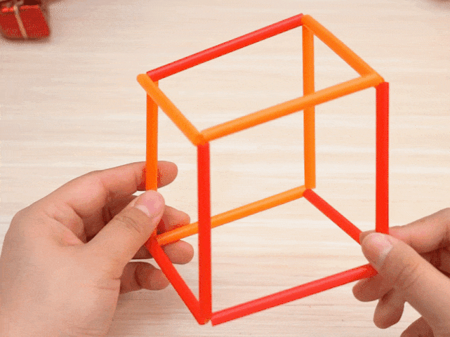 Two hands manipulate a structure made of red and orange plastic straws. The straws are slotted together to form a three-dimensional shape, which is rotated and flattened to demonstrate its flexibility and the different forms it can take. The video showcases a simple construction or puzzle made from everyday materials.