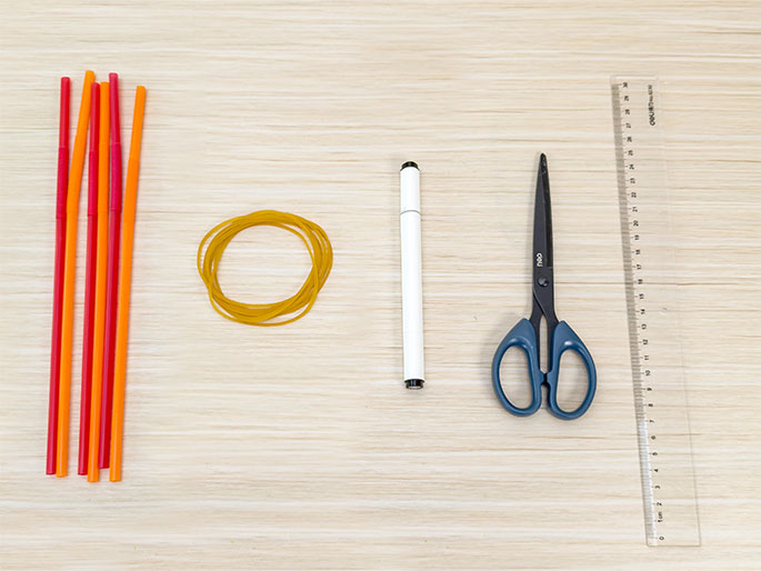 Several items are arranged on a light wood-grain surface, likely for a craft or science project. These include a small bundle of red and orange drinking straws, a yellow rubber band, a white marker pen, a pair of blue-handled scissors, and a clear plastic ruler. The arrangement suggests preparation for a hands-on activity.