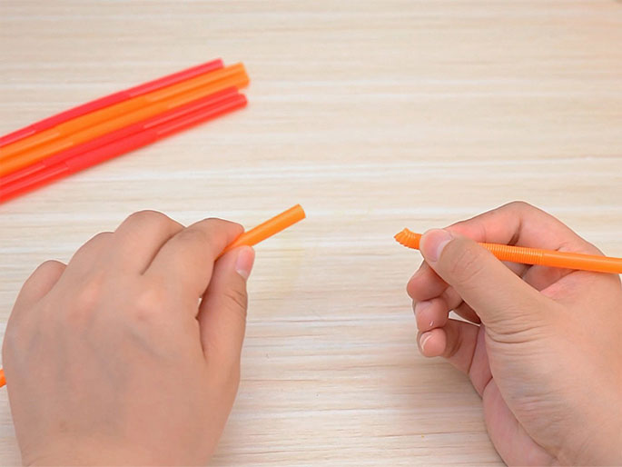 Two hands manipulate sections of orange plastic drinking straws against a light wood-grain background. One hand holds a full, uncut piece of straw, while the other holds a shorter piece with a diagonal cut at one end, revealing the corrugated inner structure. A small pile of uncut straws is visible in the background. The image suggests a craft or DIY project involving the straws.