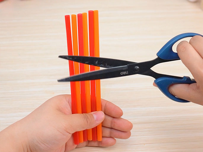 A person is using a pair of blue-handled scissors to cut a bundle of red and orange drinking straws. One hand holds the straws together while the other operates the scissors. The backdrop is a light wood-grain surface. The image conveys a craft or DIY activity involving cutting the straws.