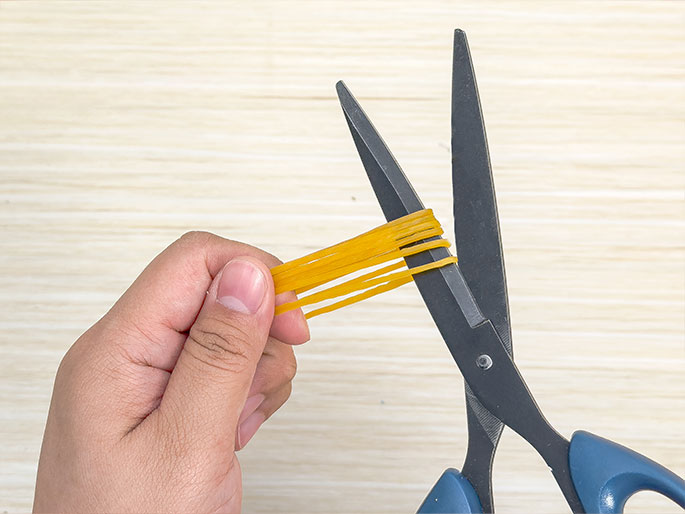 A hand holds a cut rubber band stretched taut, with numerous thin strands splayed out like spaghetti. A pair of blue-handled scissors is positioned as if in mid-cut, having just created the many strands from the original rubber band. The background is a light wood-grain surface. The image suggests a DIY or craft project, or possibly an experiment involving the properties of rubber bands.