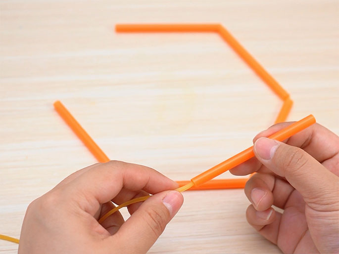 Two hands work on threading a thin, light-yellow rubber band through short segments of orange drinking straws. A partially constructed hexagonal shape made of connected straw segments is visible on the light wood-grain background. The hands are in the process of adding another straw piece to the structure using the rubber band. The image suggests a craft or construction activity.