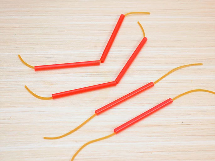 Several short segments of red drinking straws are laid out on a light wood-grain surface, connected by lengths of thin, amber-colored rubber bands. The straws and rubber bands form a loose, abstract pattern, with some sections resembling connected joints or limbs. The image suggests components for a craft project or a deconstructed version of a previously assembled object.