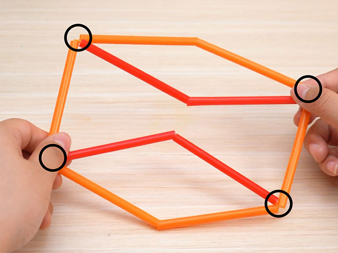 Two hands hold a geometric shape constructed from short segments of orange and red drinking straws. The shape resembles an irregular hexagon or a flattened cube, with the straws forming the edges. The points where the straws are connected are clearly visible. The structure rests on a light wood-grain background. The image suggests a craft project or a geometric model made from simple materials. The circled areas highlight the straw connections, emphasizing the construction technique.
