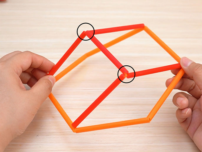 Two hands hold a three-dimensional structure made of short orange and red drinking straw segments. The structure resembles a simplified cube or rectangular prism, viewed at an angle. The connections between the straws are clearly visible, and are highlighted by black circles. The object sits on a light wood-grain background. The image suggests a craft project, a geometric model, or a simple construction made for educational or recreational purposes.