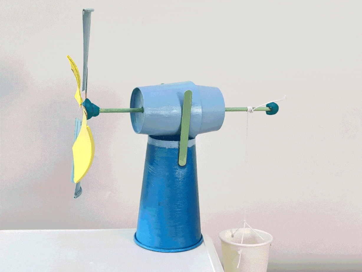 A small windmill model made from paper cups, craft sticks, and paper spins on a white table. A miniature bucket hangs from an arm attached to the windmill.