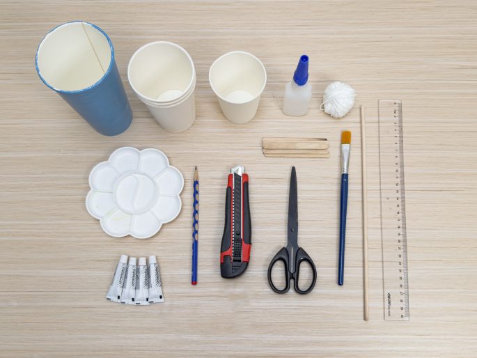 An overhead flat lay showcasing a collection of craft supplies arranged on a light wood-grain table. The supplies include a blue painted paper cup, several white paper cups, a bottle of white glue, a ball of white yarn, a ruler, wooden craft sticks, a paintbrush, scissors, a utility knife, a pencil, a flower-shaped paint palette, and several small tubes of paint or glue. These materials appear to be gathered for a crafting project.