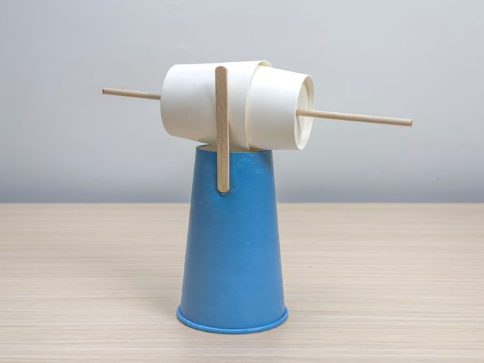 A simple craft resembling a miniature windmill or seesaw sits on a light wood-grain table. A small, light-blue paper cup serves as the base. Two thin wooden dowels are inserted into the cup, and a flattened white paper cup is attached to the dowels, balancing horizontally. This image showcases a finished DIY craft project.