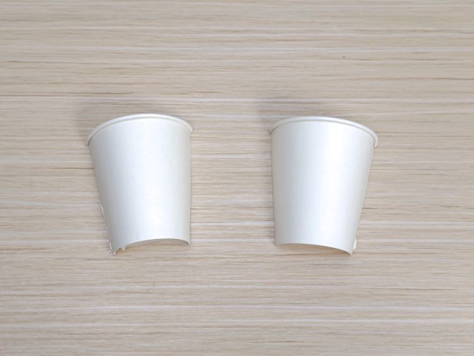 Two identical white paper cups sit on a light wood-grain surface. Both cups have a section cut out from their sides, making them appear partially flattened. This image seems to depict a step in preparing materials for a craft project.