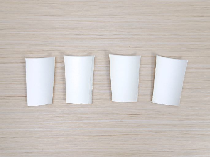 Four identical white paper cups are arranged in a row on a light wood-grain surface. Each cup has a section cut out from the side, giving them a partially flattened appearance. These appear to be prepared materials for a craft project.