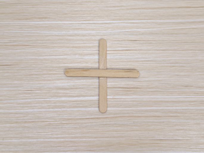 Two small, light-colored wooden craft sticks are arranged in a cross shape on a light wood-grain surface. One stick lies horizontally, and the other vertically, overlapping in the center. This image appears to show a simple craft component.