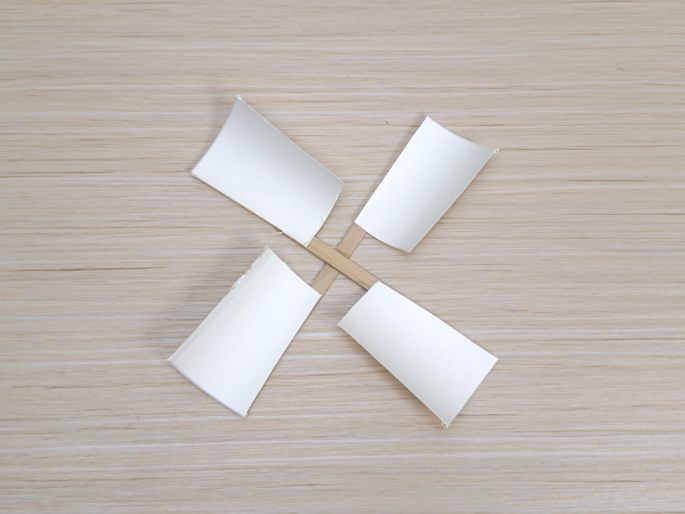 Four flattened, white paper cup pieces are attached to two small, crossed wooden craft sticks, forming a pinwheel shape on a light wood-grain surface. This image appears to show another stage in a craft project.