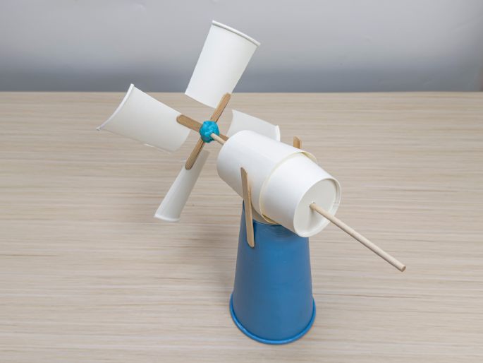 A small, homemade windmill craft sits on a light wood-grain table. The windmill's base is a light-blue paper cup. A flattened white paper cup is attached to the base with wooden craft sticks, acting as a fulcrum. A longer wooden dowel extends horizontally from this cup. At the other end of the dowel, four flattened white paper cup pieces are attached to crossed craft sticks, forming the windmill's blades. A small ball of blue putty or clay secures the blades to the dowel. This image showcases a completed DIY windmill project.