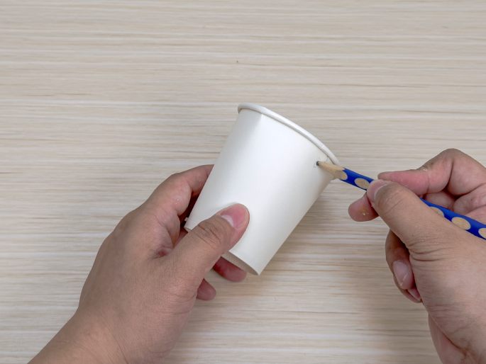 Two hands hold a white paper cup over a light wood-grain surface. One hand holds the cup steady, while the other uses a short, sharpened pencil to widen a small hole near the cup's rim. This image appears to depict a step in a craft project involving repurposing a paper cup.