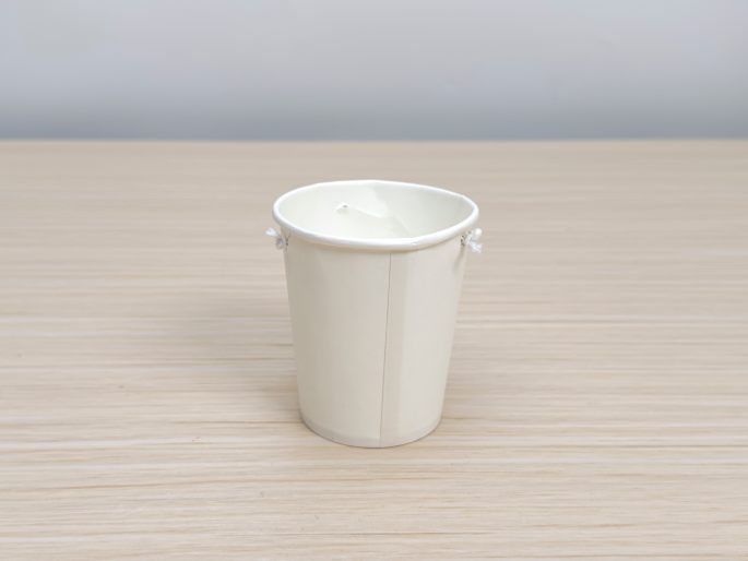 A small, white paper cup sits on a light wood-grain table. Short lengths of string are tied through small holes on opposite sides of the cup's rim, creating a small, improvised bucket or pail. This appears to be another element for a craft activity.