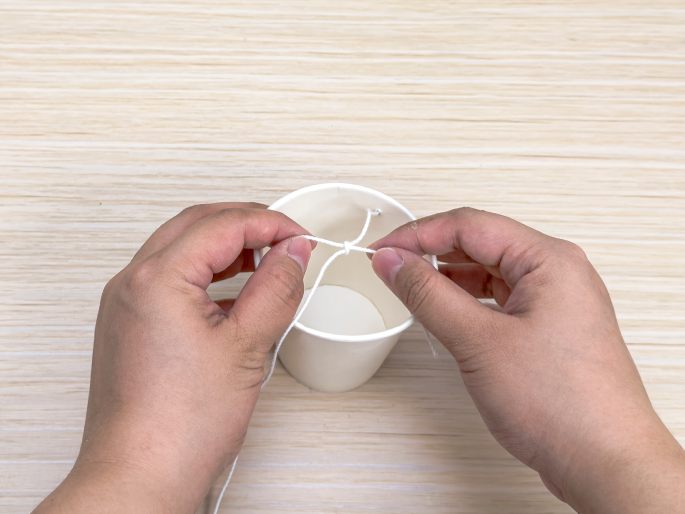 Two hands hold a white paper cup with pieces of string threaded through holes on opposite sides of the rim. The hands are in the process of tying the two strings together above the cup's opening, creating a handle. The cup sits on a light wood-grain surface. This image appears to be another step in a simple craft project.
