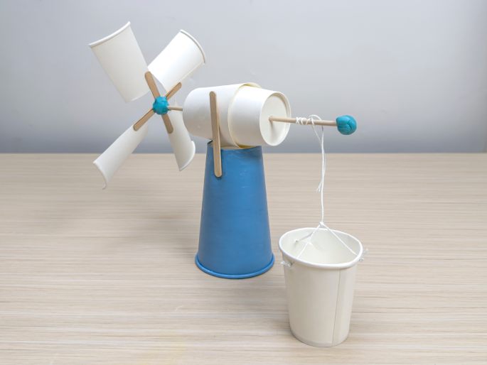 A completed, homemade windmill craft project stands on a light wood-grain table. The windmill's base is a light-blue paper cup. A flattened white paper cup is attached to the base with wooden craft sticks, acting as a fulcrum. A longer wooden dowel extends horizontally from this cup. A small ball of blue putty or clay is affixed to the dowel's end. A string is tied to this blue putty and hangs down to a small, white paper cup bucket which has string handles. At the other end of the dowel, four flattened white paper cup pieces form the windmill's blades and are attached to crossed craft sticks and held to the dowel with a small ball of blue putty. This image demonstrates a finished DIY windmill model with a small bucket suspended from it.