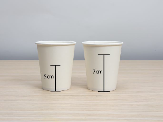 Two white paper cups sit on a light wood-grain surface. A vertical line beside each cup indicates its height. The smaller cup on the left is 5cm tall, and the larger cup on the right is 7cm tall. This image illustrates the size difference between the two cups.