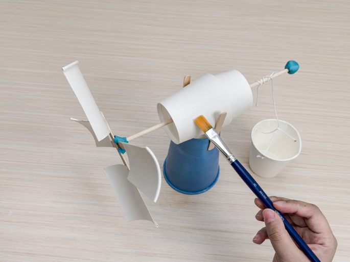 A partially constructed windmill craft project sits on a light wood-grain table. The windmill's base is a light-blue paper cup. A flattened white paper cup is attached to the base with wooden craft sticks, acting as a fulcrum. A longer wooden dowel extends horizontally from this cup. A small ball of blue putty or clay is affixed to the dowel's end. A string is tied to this blue putty and hangs down to a small, white paper cup bucket which has string handles. At the other end of the dowel, four flattened white paper cup pieces are attached to crossed craft sticks to form the windmill's blades. These blades are secured with small pieces of blue tape. A hand holds a paintbrush and is applying something to the flattened white cup that serves as the fulcrum. This image captures a stage in the creation of a DIY windmill model.