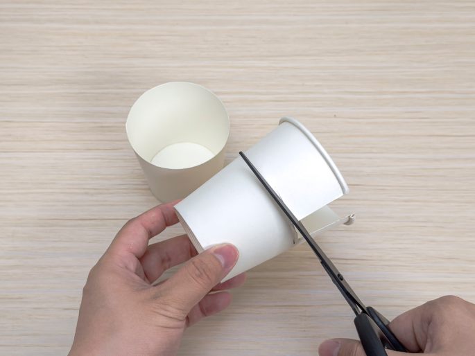 Two hands are using scissors to cut a small white paper cup. A portion of the cup has already been cut away. Another uncut paper cup sits on the light wood-grain table in the background. This image shows a step in a crafting process involving paper cups.