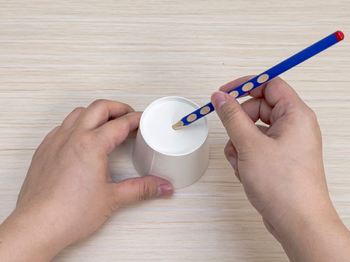 Two hands manipulate a small white paper cup and a blue pencil on a light wood-grain table. One hand holds the cup steady while the other hand uses the pencil to poke a hole in the bottom of the cup. This image depicts a step in a craft project.