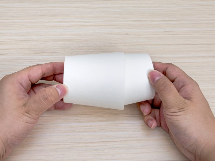 Two hands hold a flattened white paper cup against a light wood-grain background. The cup appears to have been cut and unfolded. This image likely demonstrates a step in a crafting process.