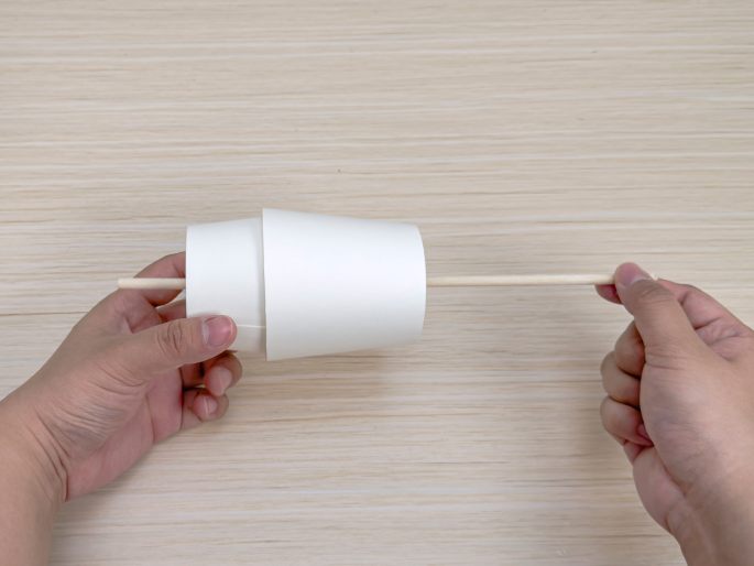 Two hands hold a flattened white paper cup and a thin wooden dowel on a light wood-grain table. The dowel is inserted through the cup. This image appears to show a stage in constructing a simple craft or toy.