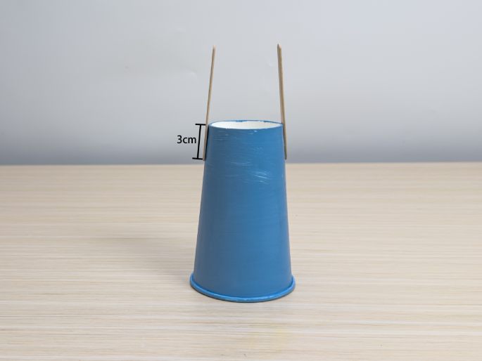 A small, light-blue paper cup sits on a light wood-grain table. Two thin, light-colored wooden dowels are inserted into the top of the cup on opposite sides, sticking upwards. A bracket and the label 3cm indicate the height of the dowel protruding from the cup. This image appears to show a component of a craft project.