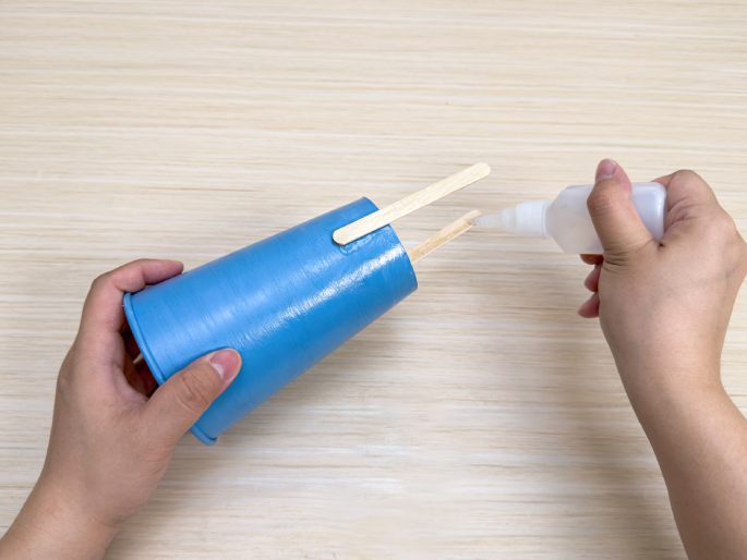 Two hands work on a craft project on a light wood-grain table. One hand holds a small light-blue paper cup with two wooden dowels inserted into the top. The other hand holds a small bottle of glue, applying it to the dowels and cup. This image shows a step in assembling a craft.