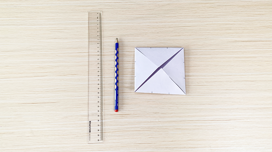 A flatlay shows a partially folded piece of white paper, a blue pencil, and a ruler on a light wood surface, suggesting the tools and materials for a paper craft project.