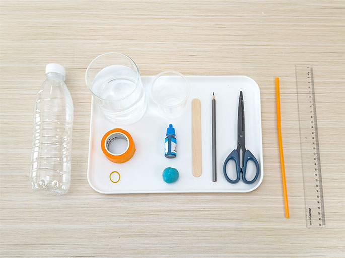 An assortment of materials arranged on a light wood-grain table appears ready for a science experiment or craft project. A white rectangular tray holds two clear glass containers, one partially filled with water, a roll of orange tape, a small bottle of blue liquid, a lump of blue putty or clay, a wooden craft stick, a pencil, and a pair of scissors. A clear plastic water bottle lies beside the tray, and an orange drinking straw and a clear plastic ruler are positioned next to the tray. The image suggests preparation for a hands-on activity.