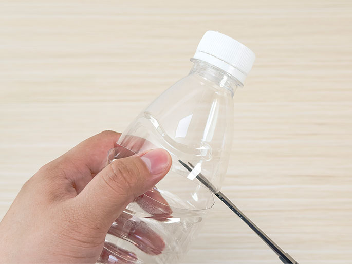 A hand holds a clear plastic water bottle against a light wood-grain background. A pair of scissors is positioned to cut the bottle near its base. The image suggests the bottle is being repurposed or modified for a craft project or experiment.