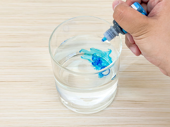 A hand holds a small bottle of blue dye or ink over a clear glass of water, set on a light wood-grain background. Drops of the blue liquid are being dispensed into the water, creating swirling patterns and demonstrating the diffusion or mixing of liquids.