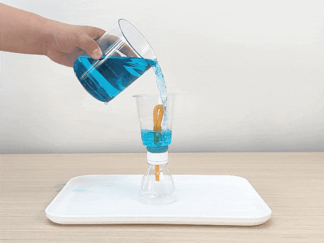A hand pours bright blue liquid from a clear beaker into a clear plastic cup positioned on top of an inverted clear plastic bottle on a white tray. The bottle and cup are connected by an orange straw sealed with turquoise putty. The blue liquid is partially filling the cup, and the setup suggests a science experiment demonstrating air pressure, siphoning, or fluid dynamics. The background is plain white.